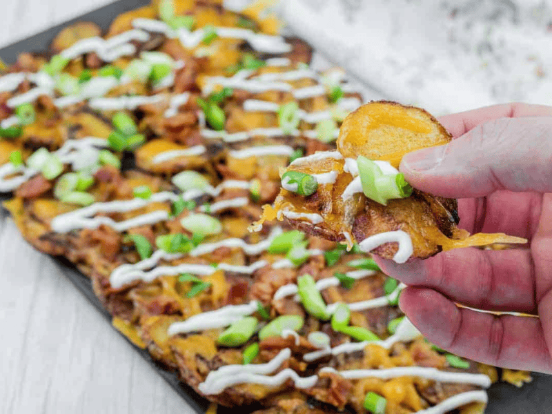 Hand holding a loaded Irish nachos with cheese, bacon, sour cream, and green onions, over a platter of similarly topped slices on a table.