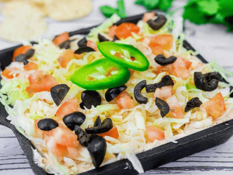 A skillet of Taco dip filled with shredded lettuce, diced tomatoes, sliced black olives, and jalapeño slices, served on a light wooden surface.