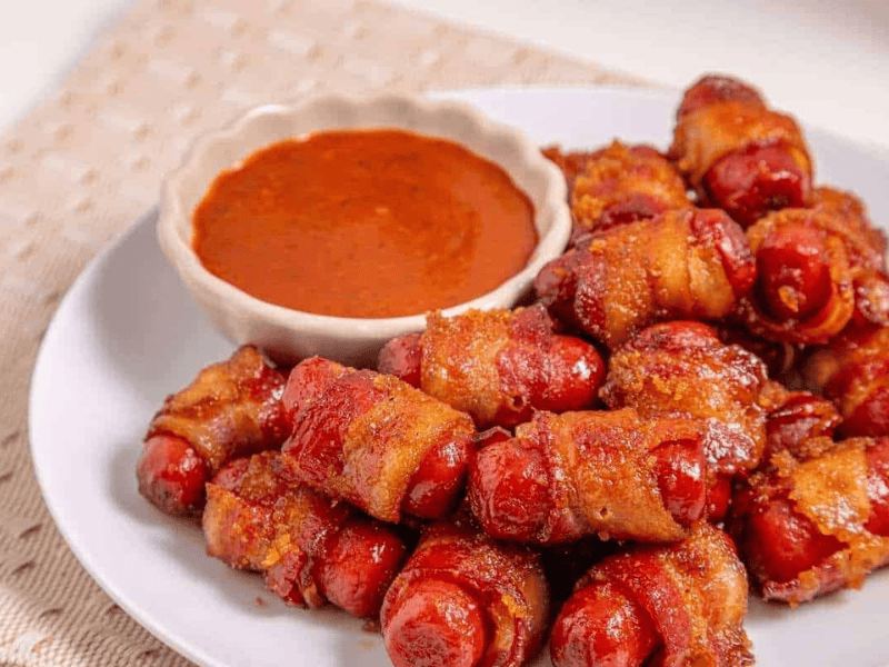 A plate of bacon-wrapped smokies sits next to a small bowl of reddish dipping sauce on a light-colored surface.