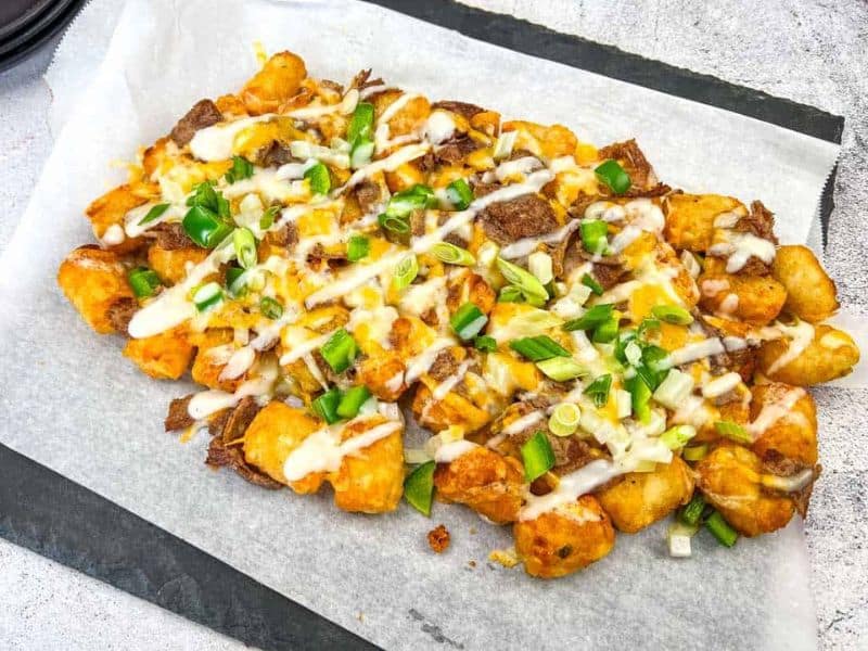 Philly Cheesesteak Loaded Tots on parchment paper, topped with Alfredo cheese, green onions, and a drizzle of white sauce, placed on a dark rectangular slate plate.